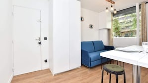 A compact living space with a white wardrobe, a blue sofa, a dining table with a stool, and a window with a view of buildings. - Modern 2BD apartment in Parc Belleville (Paris)