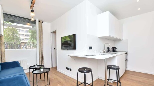 A modern apartment with a kitchenette, a sofa, two bar stools, and a TV mounted on the wall. - Modern 2BD apartment in Parc Belleville (Paris)