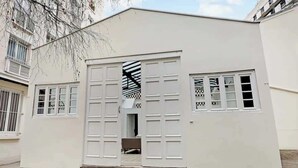 Un bâtiment blanc doté d’un grand portail de garage, d’une fenêtre à motifs à carreaux et d’une porte donnant sur un espace intérieur.