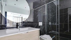 Une salle de bain moderne avec un mur de douche en marbre, un grand miroir et un lavabo blanc.