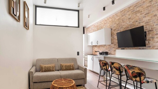 A modern living space with a sofa, a dining area with chairs, a TV mounted on a brick wall, and a kitchen in the background. - 4 Chambres, 4 Salles de Bains Proche Gare de Lyon (Paris)