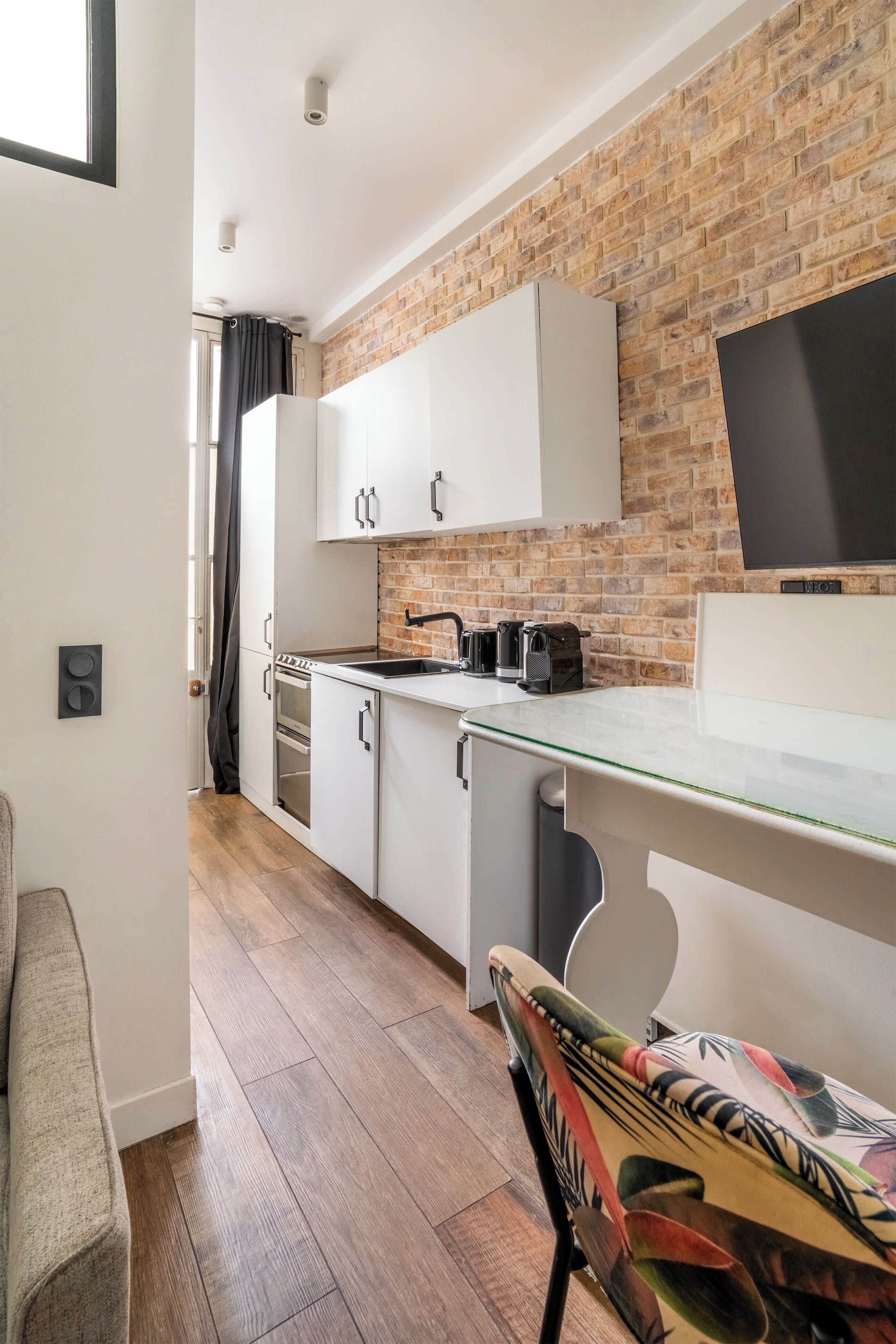 A compact kitchen with white cabinets, a brick wall, a flat-screen TV, and a wooden floor.