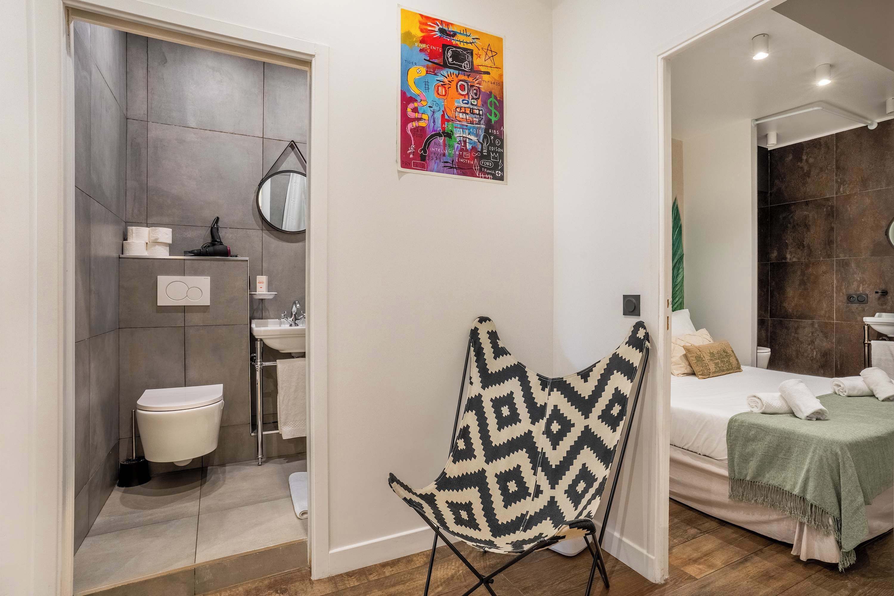 A modern bathroom with a toilet, sink, and shower. There is a colorful abstract painting on the wall and a patterned chair in the room.