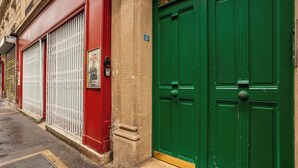 Vista da rua com um prédio vermelho e portas verdes, uma placa com o número '35' e uma grade de metal na calçada.