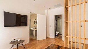 Quarto de hotel moderno com televisão de tela plana, uma pequena mesa redonda, piso de madeira e uma porta deslizante para o banheiro.