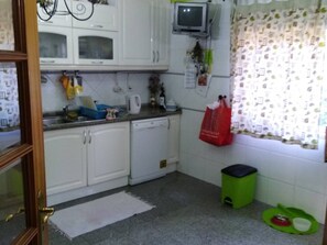 Uma cozinha com armários brancos, micro-ondas, televisão pequena, lixeira verde e uma janela com cortinas estampadas.