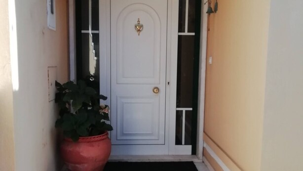 Una porta bianca con una basetta di ottone, un tappetino nero all'ingresso e una pianta in vaso di grandi dimensioni a sinistra.