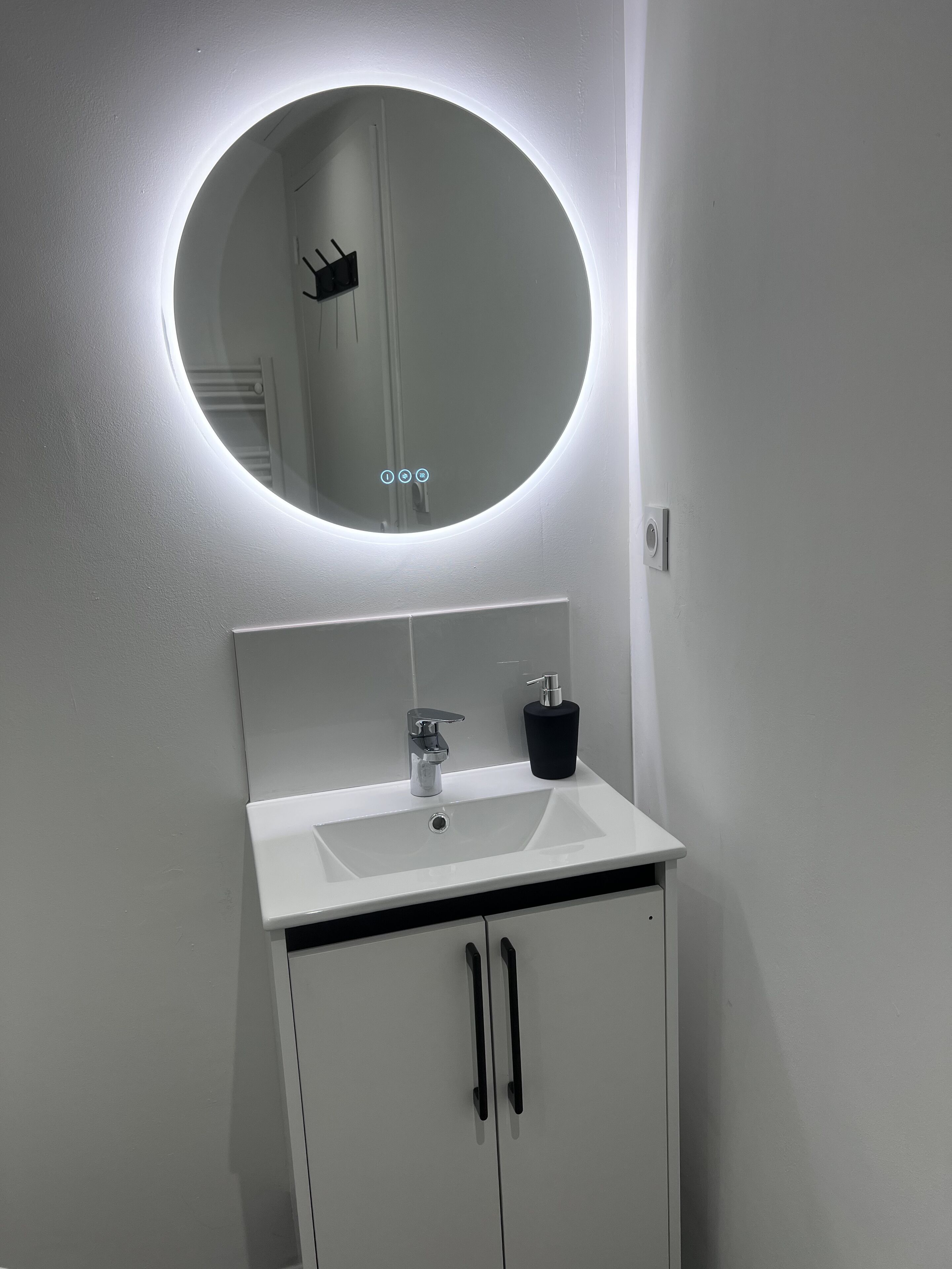 A bathroom with a white sink, a round mirror with a light, and a black soap dispenser.