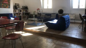 A living room with a sofa, a coffee table, a bookshelf, and a dining area with chairs and a table. - Au Cœur de Saint Germain ! 80m2 Confortable, Calme et Lumineux Avec vue (Paris)