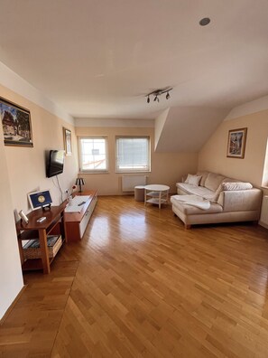 A spacious living room with a wooden floor, a sofa, a TV, and a desk with a computer.