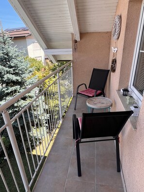 Ein Balkon mit einem schwarzen Stuhl und einem kleinen runden Tisch, einem weißen Geländer sowie Blick auf Bäume und ein Gebäude.
