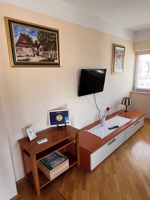 Ein Zimmer mit Schreibtisch aus Holz, einem an der Wand befestigten Fernseher, einem gerahmten Bild, einer Lampe und einem Fenster.