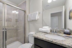 Een badkamer met een douchecabine van glas, een wit toilet, een wastafel met een granieten aanrechtblad en een spiegel.
