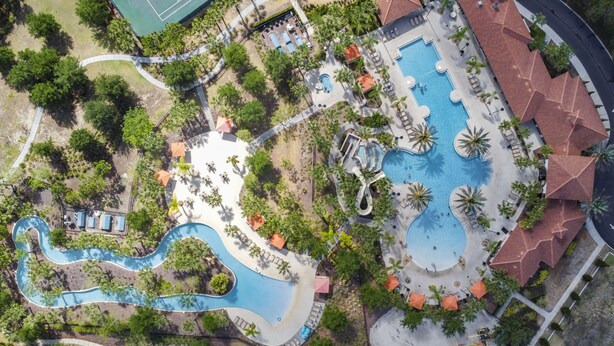 Luchtbeeld van een resort met meerdere zwembaden, een tennisbaan en weelderige begroeiing.
