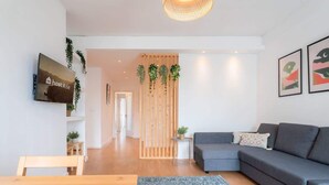 A modern living room with a grey sofa, wooden coffee table, and a blue patterned rug. A television is mounted on the wall, and there are framed artworks on the wall.