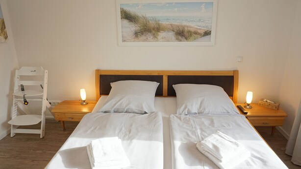 A double bed with white linens, two bedside tables with lamps, a wooden nightstand, a white chair, and a painting of a beach scene on the wall. - Ruhige Ferienwohnung in Tönning für bis zu 4 Personen (Tönning)