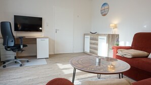 A modern living room with a flat-screen TV, a desk with a chair, a red sofa, a small round coffee table, and a wall clock. - Ruhige Ferienwohnung in Tönning für bis zu 4 Personen (Tönning)