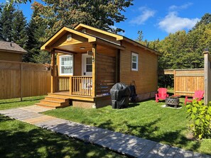 A small wooden cabin with a porch, a barbecue grill, and a wooden deck leading to the entrance. - Quaco Cottages - The William Carson (West Quaco)