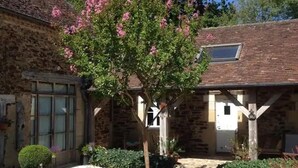 A stone building with a garden featuring a tree with pink flowers, a circular hedge, and a gravel path.