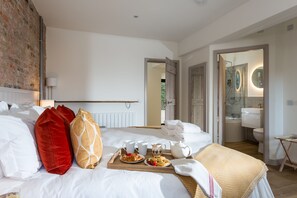 Ein Hotelzimmer mit einem aufgeräumten Bett, einem Frühstückswagen mit Gebäck und Kaffee sowie einem Badezimmer im modernen Stil.