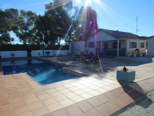 A swimming pool with a tiled deck, outdoor furniture, and a house with a white exterior and a red roof.