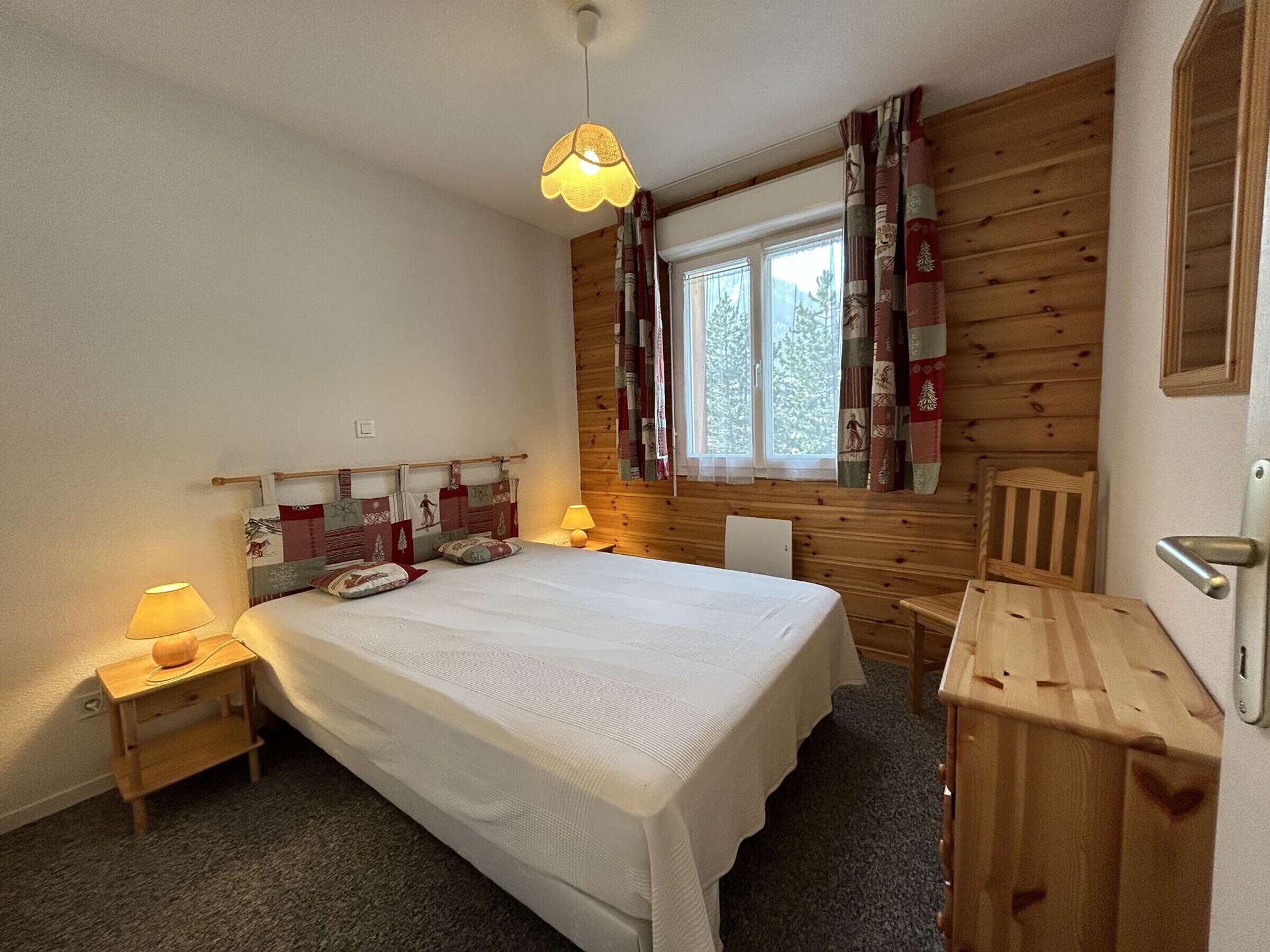 Ein Schlafzimmer mit einem großen Bett, einem Holzkopfende, einem Fenster mit Vorhängen, einem Nachttisch mit Lampe und einem Holzbeistelltisch.