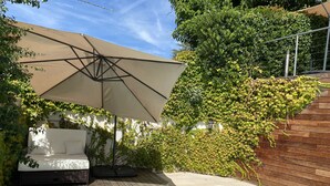 Zona junto a la piscina con terraza de madera, sofá exterior blanco y una gran sombrilla que da sombra.