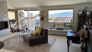 Una sala de estar con un sofá, una mesa de centro y un televisor montado en la pared. Se ve un ventanal con vistas a la ciudad y a las montañas.