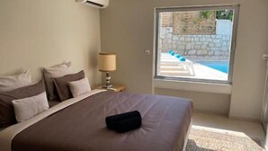 Un dormitorio con una cama grande, una mesita de noche con lámpara y vistas a una piscina a través de una ventana.