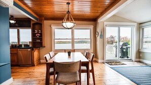 A dining area with a wooden table and chairs, a chandelier, and a view of a body of water through large windows. - Escape at the Lake: Hot tub on Lake Charlevoix (East Jordan)