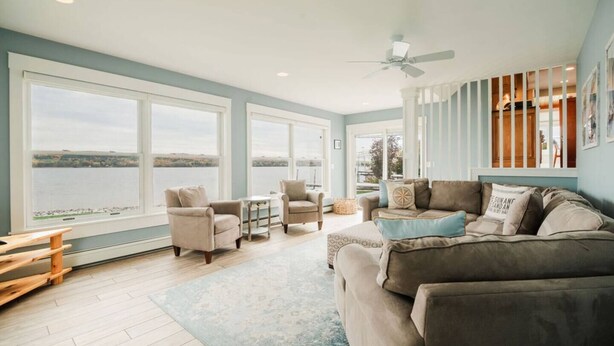 A spacious living room with large windows, a ceiling fan, and a view of a body of water. - Escape at the Lake: Hot tub on Lake Charlevoix (East Jordan)