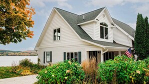 A white two-story house with a gabled roof, a covered porch, and an American flag. - Escape at the Lake: Hot tub on Lake Charlevoix (East Jordan)