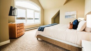 A bedroom with a large bed, a wooden dresser, a ceiling fan, a window with a view, and a painting on the wall. - Escape at the Lake: Hot tub on Lake Charlevoix (East Jordan)