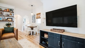 A modern living room with a flat-screen TV, a wooden coffee table, a brown sofa, and a dining area with white chairs.