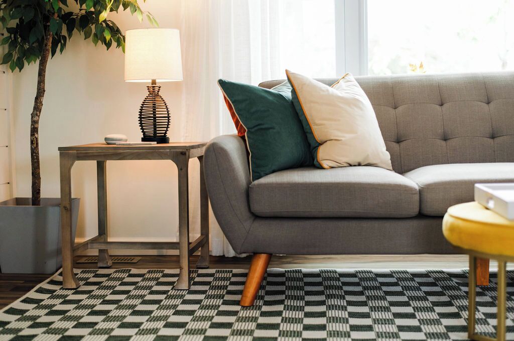A living room with a grey sofa, a patterned rug, a small table with a lamp, and a potted plant.