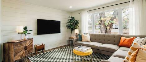 Una sala de estar con piso a cuadros, un sofá, un televisor montado en la pared y una ventana con cortinas.
