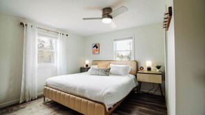 Un dormitorio con cama, ventilador de techo, dos mesitas de noche con lámparas y una ventana con cortinas.