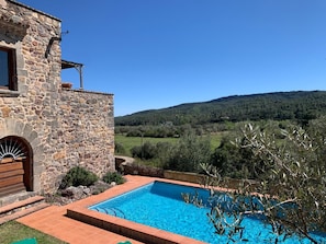 A stone building with a swimming pool, surrounded by olive trees and a view of a hilly landscape.