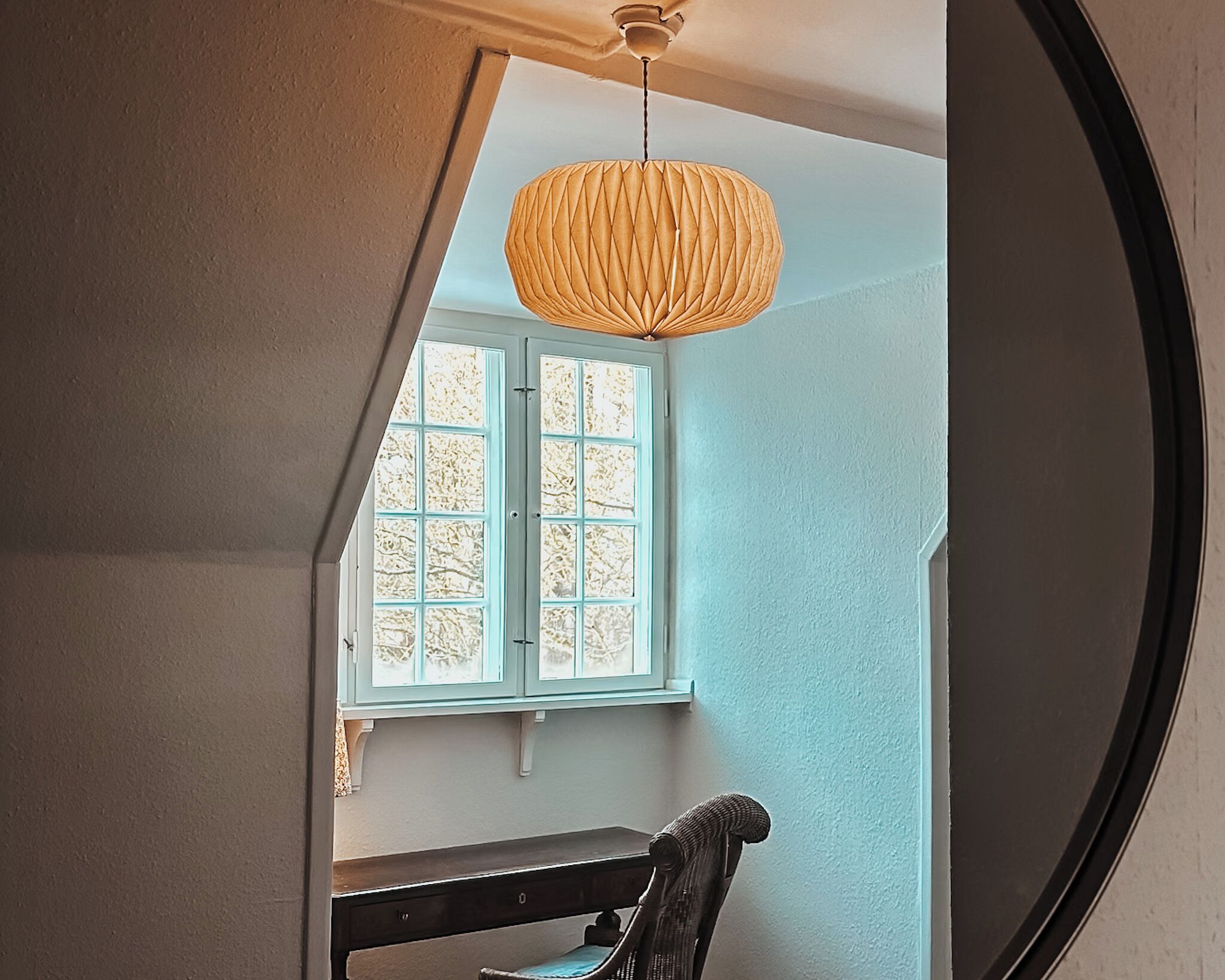 A room with a large mirror, a window with a view of trees, a wooden bench, and a hanging light fixture.