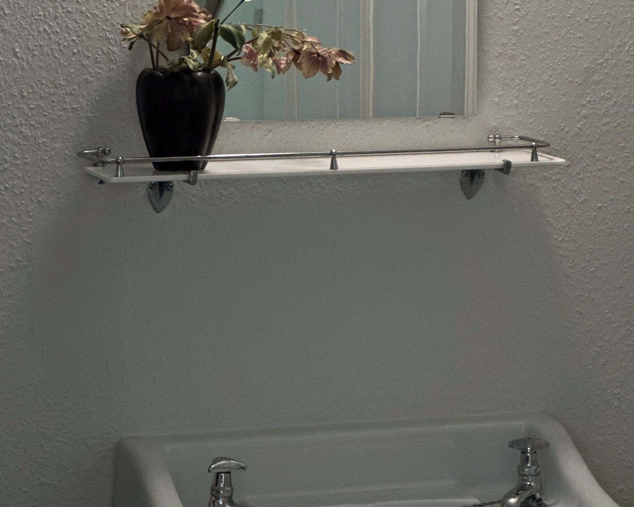Une salle de bain avec un lavabo blanc, un miroir et une étagère sur laquelle est posé un vase de fleurs.