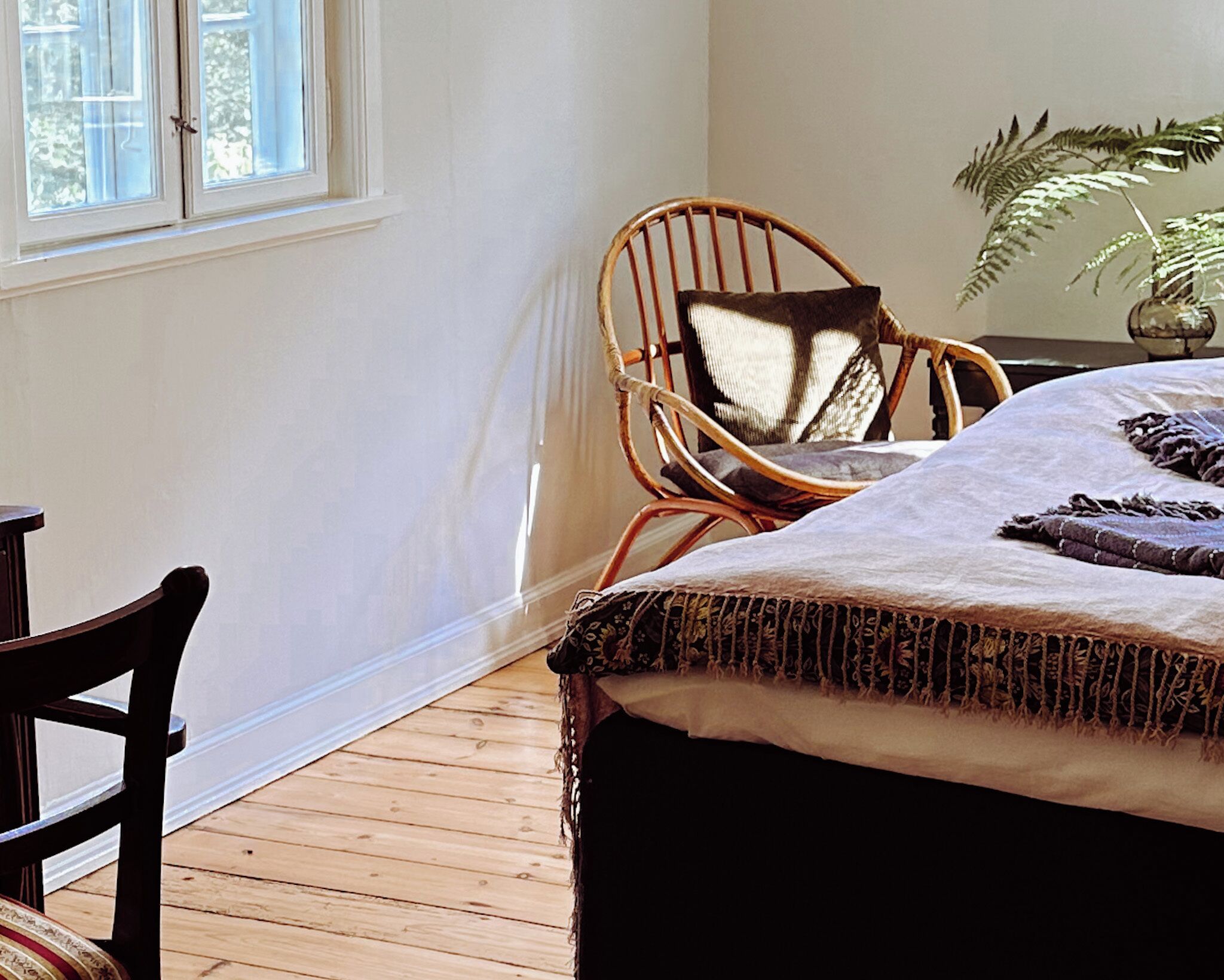 Un dormitorio con piso de madera, una cama con cubrecama estampado, una silla de mimbre con cojín y una planta en maceta.