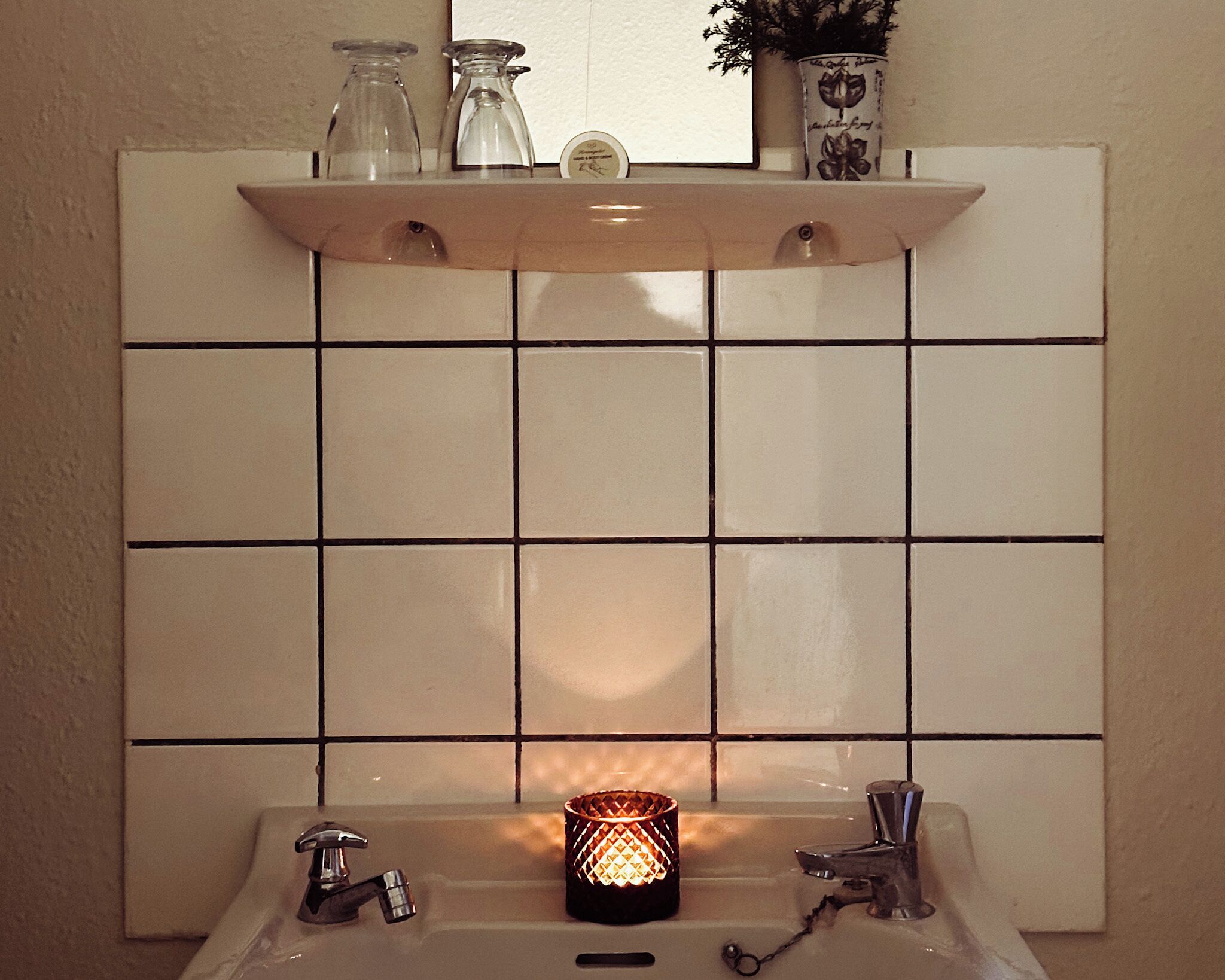 A bathroom with a tiled wall, a shelf above the sink, and a lit candle on the sink.