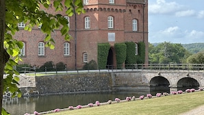 A historic brick building with a clock tower, surrounded by a moat and pink flowers. - The Woodland Lodge Gisselfeld (Haslev)