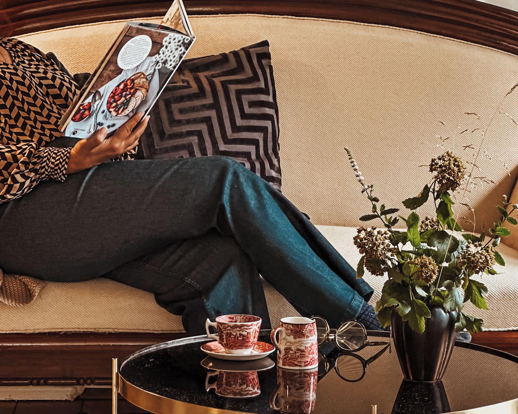 A person sitting on a sofa with a magazine, a cup, and a vase with flowers on a side table.