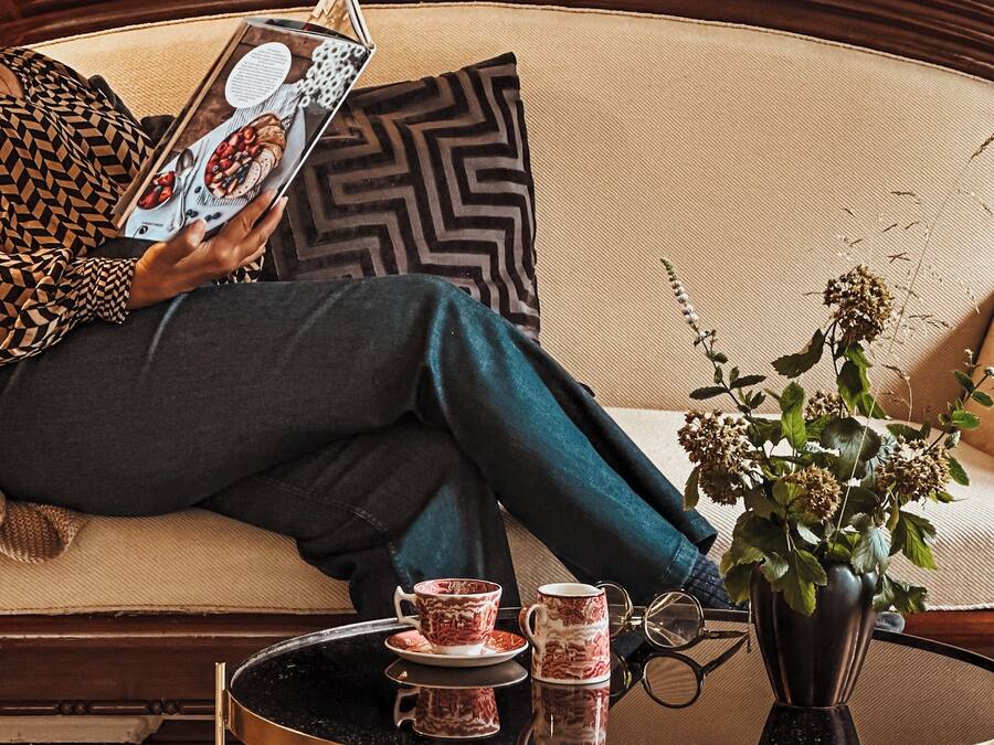 A person sitting on a sofa with a magazine, a cup, and a vase with flowers on a side table.
