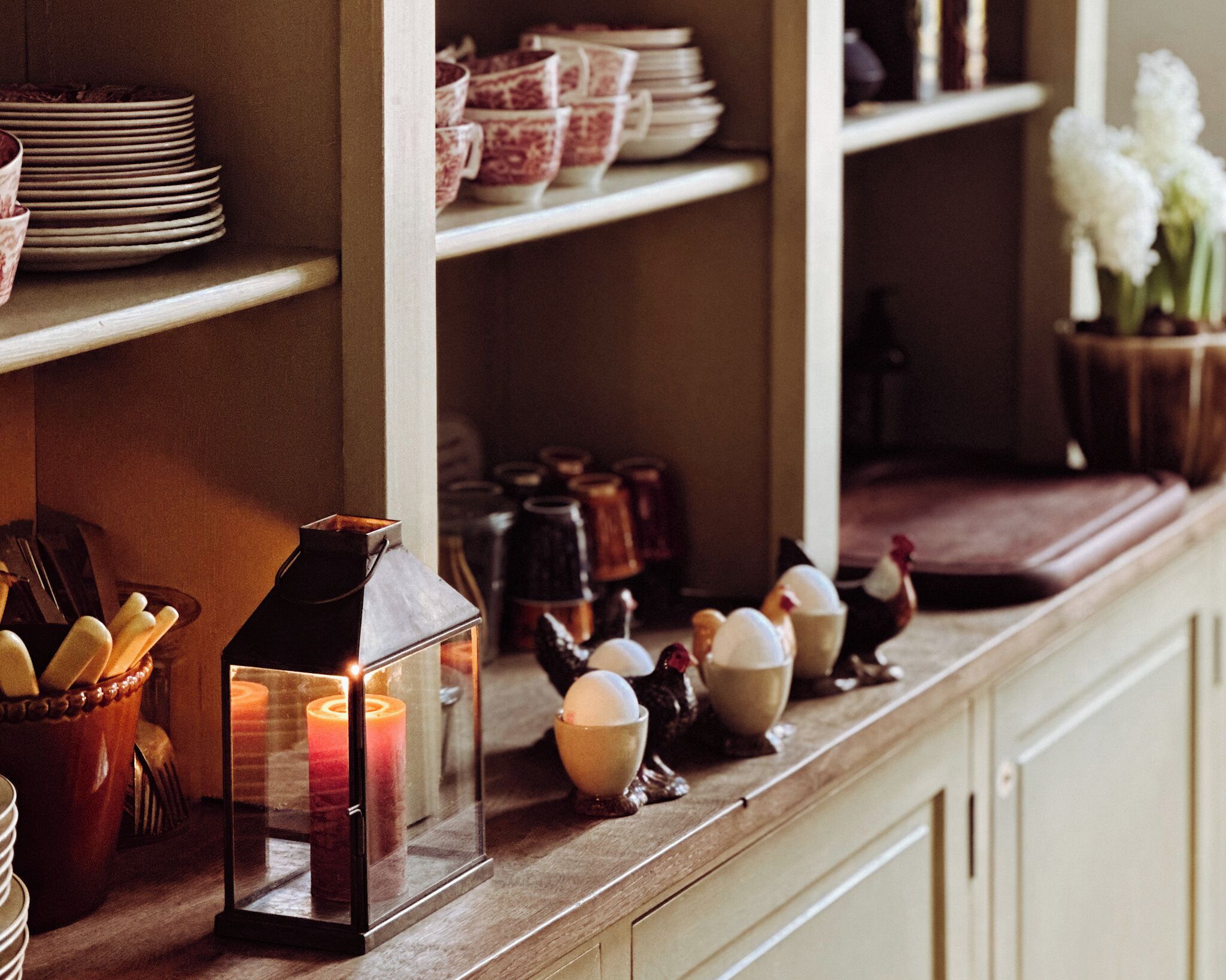 A kitchen counter with a lit candle in a glass lantern, ceramic eggs, and stacked plates.