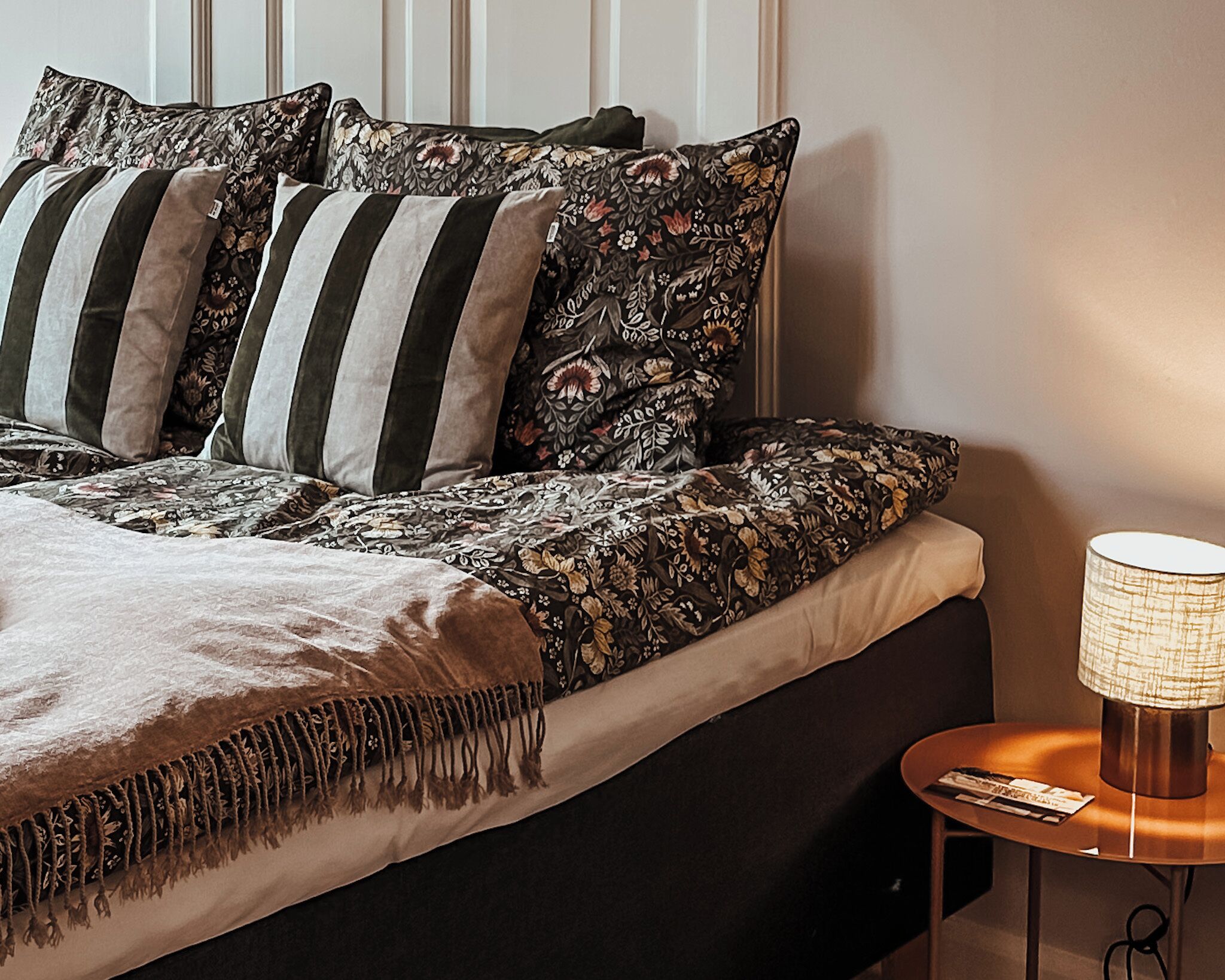 A neatly made bed with striped and floral pillows, a throw blanket, and a bedside table with a lamp.