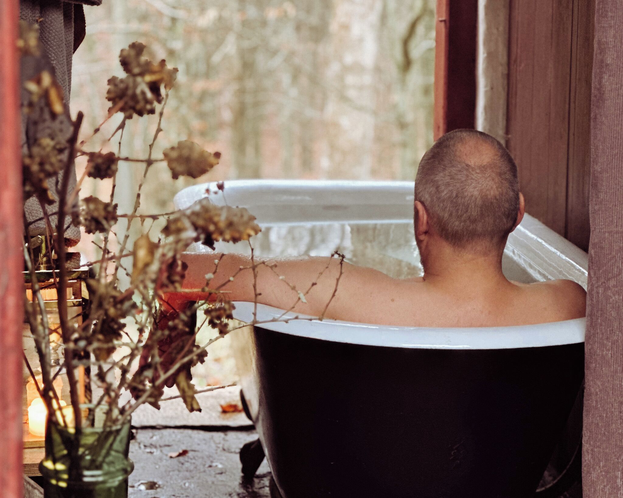 Una persona en una bañera con vista a una pared de piedra y flores secas afuera.