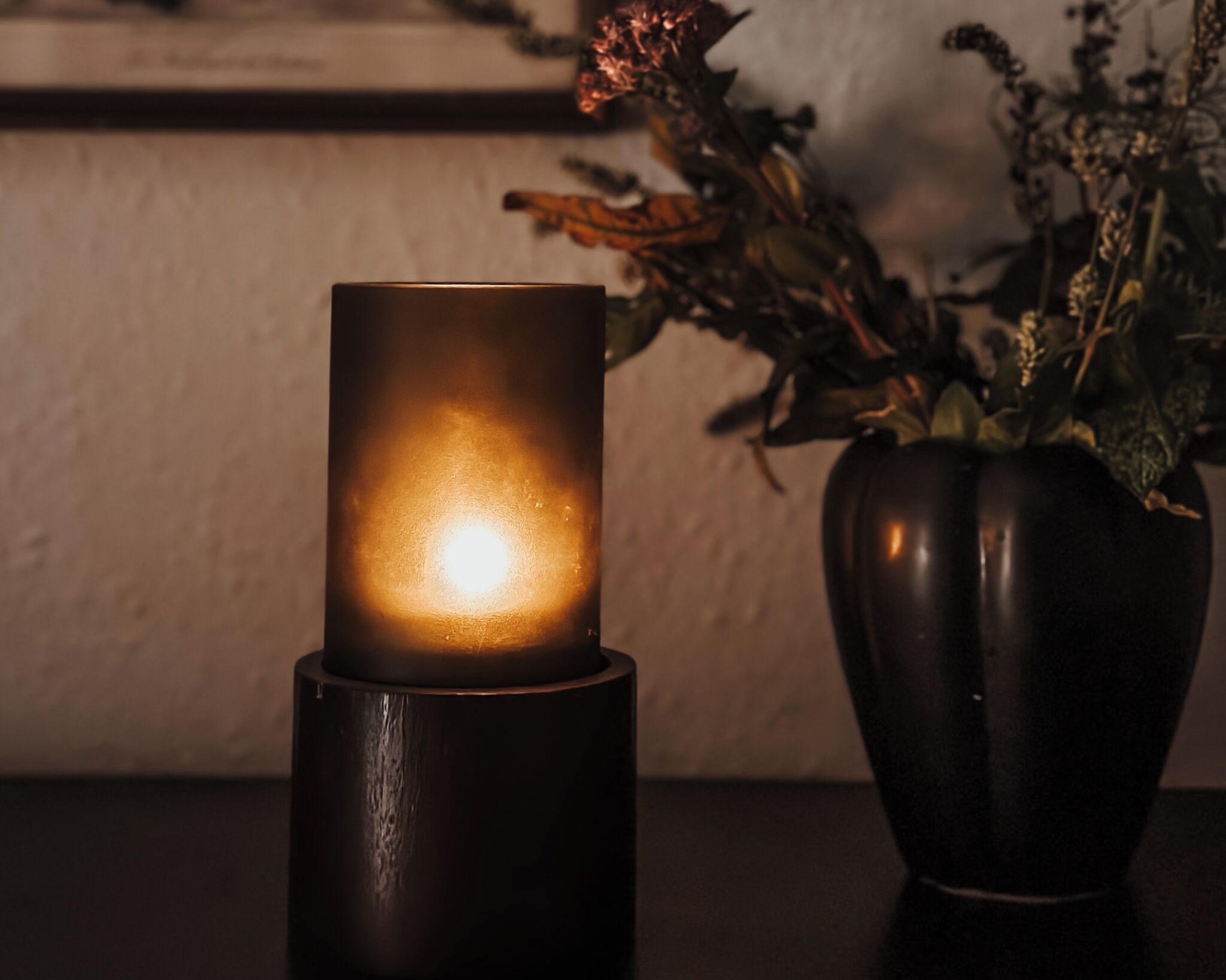 Una vela en un soporte oscuro sobre una mesa, junto a una maceta con flores secas.