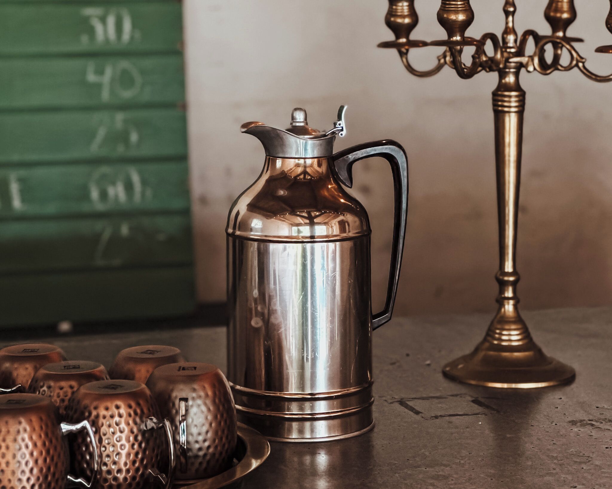 A stainless steel coffee pot on a table with a green background and a brass candelabra.